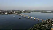 Farbfotografie in Vogelperspektive auf den Fluss mit seiner Brücke. Im Hintergrund ist die Stadt und flache Waldlandschaft zu erkennen.