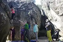 Mehrere Personen zwischen Sandsteinblöcken; eine Kletterin an einer Wand, ca. 2 m über dem Boden; ein Mann hat beide Arme in Richtung der Kletterin ausgebreitet