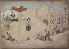 Okuni steht auf einer Bühne, vor der mehrere Zuschauer sitzen. Sie trägt einen schwarzen Kegelhut, ein beigenes Oberteil und eine weite, schwarze Hose mit weißem Pflanzenmuster. In der linken Hand hält sie einen Gong, in der rechten einen schwarzen Gongschlägel. Hinter ihr sitzen mehrere als Priester verkleidete Schauspieler. Sie tragen einen Chonmage, kahl rasiertes Haupthaar plus Pferdeschwanz. Die Priester tragen verschiedenfarbige Gewänder, einer von ihnen schlägt eine kleine Trommel.
