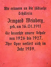 Eine Gedenktafel aus altrosa-farbenem, poliertem Stein, vielleicht Granit. Sie trägt in schwarzer Frakturschrift den Text: „Wir erinnern an die jüdische Schülerin Irmgard Weinberg, geb. am 16.01.1911. Sie besuchte unsere Schule von 1926 bis 1927. Ihre Spur verliert sich im Jahr 1939.“
