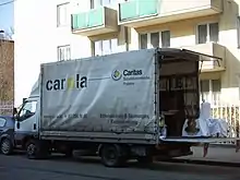 LKW mit der Aufschrift "carla" (Caritas Lager) parkt mit heruntergelassener Hebebühne vor einem Wohnhaus.