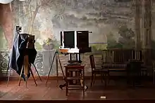 Farbfotografie von drei Balgenkameras, die vor einer Wand mit Landschaftsmalerei stehen. Die Kameras werden von links nach rechts immer größer und stehen auf Stativen. Rechts befindet sich eine Sitzgruppe.