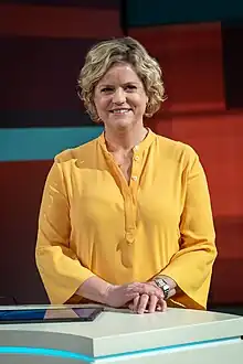 Brigitte Büscher in gelbem Oberteil vor dunkelroter Kulisse in einem Fernsehstudio