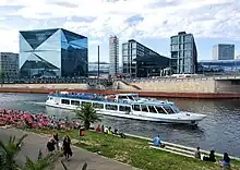 Frontale Farbfotografie auf das Glasgebäude. Direkt an der Uferpromenade stehen zwei sechs geschossige Teilgebäuden mit einer kleineren Glasfront in der Mitte, die als Aufschrift „DB Berlin Hauptbahnhof“ hat. Alle drei Gebäude sind von einer großen Glasröhre durchzogen. An der linken Seite steht ein grauer Turm mit dem DB Logo.