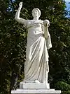 Statue von Urania im Park Sanssouci, Potsdam, Frontalansicht; ihr rechter Arm ist über ihr erhoben, die Finger zusammengebogen, eventuell war dort einmal ein Stab hineingesteckt; ihr linker Arm ist halb nach vorn ausgestreckt, in der Hand hält sie eine Kugel