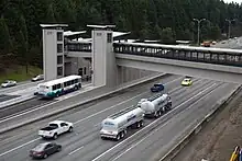 Vielspurige Autobahn mit leichtem Verkehr. Mittig eine massive Busstation mit einer über die Fahrbahn führenden, umschlossenen Fußgängerbrücke mit durchgängigem, dunkelblauem Fensterband. Ein Bus fährt gerade von links kommend ein. Farben Grau und Asphalt dominieren, hinten ein dunkelgrüner Wald.