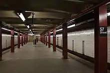 Blick entlang des Mittelbahnsteigs der Bahnhofs 57th Street. Der Raumeindruck wird bestimmt von den cremefarben gefliesten, teils schmutzigen Hintergleiswänden, den massiven dunkel-weinroten T-Trägern, die als Stützenreihen in der Nähe der beiden Bahnsteigkanten verlaufen, von den dunkel angelaufenen Betonplatten des Bahnsteigbodens und von einer dunkelbeige angestrichenen Decke aus quer verlaufenden Stahlträgern und vermutlich Betonfüllungen. Entlang der Decke an den Bahnsteigkanten verlaufen ununterbrochene Bänder von Leuchtstoffröhren, von denen aber manche defekt sind und nicht leuchten. In der Bahnsteigmitte ist eine dritte, allerdings aus Einzelröhren mit jeweils Lücken zwischen den Leuchtstofflampenhaltern bestehende Lichtreihe. Im Bildhintergrund blickt man direkt auf zwei jeweils die halbe Bahnsteigbreite einnehmende Treppen, die vom Bahnsteig nach oben führen. An jeder zweiten der dunkelroten Bahnsteigstützen sind Schilder mit dem Bahnhofsnamen montiert im typischen Transit-Authority-Design in der Schreibweise „57 Street“ mit weißer Schrift auf schwarzem Grund. An den Hintergleiswänden ist der Bahnhofsname in regelmäßigen Abständen aufgedruckt in der Schreibweise „57th Street“.