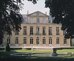 Blick aus dem Garten über ein kleines Wasserbassin auf die dreigeschossige Fassade des Hôtels de Castries mit vielen hohen Fenstern und einem mittig positionierten Dreiecksgiebel auf Dachgeschosshöhe. Über die gesamte Breite ist dem Gebäude eine Terrasse vorgelagert, zu der eine breite Treppe mit nur wenigen Stufen hinaufführt. Auf der Terrassenbrüstung stehen weiße Steinvasen. Am rechten und linken Bildrand sind dicht belaubte Bäume zu sehen.