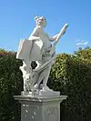 Statue von Klio im Park Belvedere, Wien, Schrägansicht von vorn links; Klio stützt sich mit der rechten Seite auf ein übergroßes Buch, das auf einem Baumstumpf steht, in der linken Hand hält sie ein Blasrohr