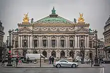 Frontale Farbfotografie eines zweigeschossigen Gebäudes im Neobarock-Stil. Im Erdgeschoss sind Rundbögen und im Obergeschoss sind zwischen den Fenstern Säulen, über denen „Académie nationale de musique“ steht. An den Dachecken stehen zwei goldene Figuren und das Gesims ist Gold. In der Mitte ist eine grüne Kuppel mit Figuren, die eine goldene Krone hochhalten.