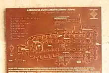 Eine rostbraune Metalltafel. Der Grundriss der Kathedrale ist als Relief erhaben und somit auch tastbar dargestellt, die Beschriftung ist in Normalschrift (weiß auf rostbraunem Grund) und in Brailleschrift ausgeführt.