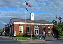Ein niedriges Backsteingebäude mit einer quadratischen Kuppel in der Mitte des mit Kupfer gedeckten Daches. Die Inschrift an der Vorderseite des Gebäudes lautet UNITED STATES POST OFFICE und ALBION NEW YORK. Eine amerikanische Flagge weht am Flaggenmast vor dem Gebäude