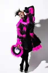 Porträtfoto von Alex Newell, der vor einer weißen Wand steht. Er hat das linke Auge geschlossen und das rechte geöffnet. Er trägt rosa Lippenstift, blauen Lidschatten, Wimperntusche, ein schwarzes Oberteil mit schwarzen und pinken Plüschstellen, schwarze Strümpfe und schwarze High Heels. In der rechten Hand hält er einen kleinen schwarzen Stoffring mit pinker Plüschumrandung. Die linke Hand hält sich Newell an den Kopf. Seine schwarzen Haare trägt er als nach oben abstehende, gekräuselte Frisur.