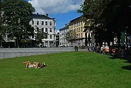 Foto einer Wiese mit sich sonnenden Personen, rechts davon eine Bewirtungsstätte und gegenüber städtische Bebauung