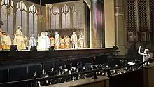 Im Hintergrund eine Opernbühne mit Mitwirkenden, im Vordergrund ein Orchester im Orchestergraben, dirigiert von Alma Deutscher.