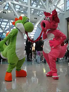 Das Foto zeigt zwei Cosplayer auf einer Messe, die als Yoshi und Birdo verkleidet sind und zueinander gerichtet stehen. Yoshi ist ein grüner, anthropomorpher Dinosaurier mit weißem Bauch und großer, runder Nase. Birdo ist ein pinker, anthropomorpher Dinosaurier mit weißen Bauch, großem, rundem Mund und rote Schleife auf dem Kopf. Im Hintergrund sind weitere Menschen zu sehen.