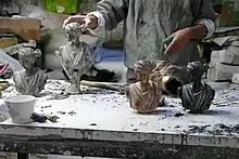 Auf einer Werkbank stehen mehrer Versionen eines Kopfes einer antiken Frauenstatue - einmal mit rötlicher Färbung, einmal schwarz und zwei graue Versionen. Dahinter der Oberkörper eines Mannes in einem grauen Pulli, der gerade eine der grauen Köpfe mit der rechten Hand berührt.