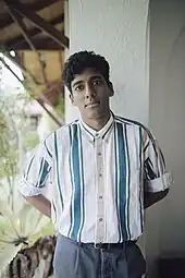 Anuk Arudpragasam ist mit hinter dem Rücken verschränkten Armen in die Kamera blickend abgebildet. Er hat kurzes schwarzes Haar und trägt ein helles Hemd das senkrechte grüne Streifen hat.
