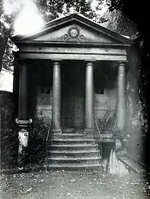 Auf der schwarz-weiß Fotografie im Hochformat ist ein Tempel zu sehen. Er hat vier Säulen und mittig ist eine Treppe. Das Dach ist in dreieckiger Form.