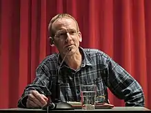 Wolfgang Berends an einem Tisch sithend mit Wasserglas und Mikrofon vor sich