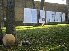 Biografietafeln sind auf einer Schnur zwischen Bäumen hängend zu sehen. Links im Bild liegt die gedenkkugel, im Hintergrund ist die Außenmauer des Lagers zu sehen.