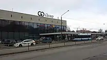 Seitliche Farbfotografie von einem Gebäude mit großer Glasfront und Reklame an dessen rechter Seite. Im oberen Bereich ist eine helle Steinfassade und auf der rechten Dachseite stehen große Reklamebuchstaben. Vom Eingang geht bis zum rechten Bildrand eine Überdachung, vor der ein blau-weißer Bus steht. Links parken einige Autos und zwei Autos fahren im Vordergrund auf der Straße.
