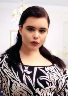 Barbie Ferreira trägt dunkelroten Lippenstift, ein schwarzes Oberteil mit weißen Blättern und Blüten sowie ihre braunen, schulterlangen Haare frei. Im Hintergrund sind ein Kronleuchter und ein eingerahmtes Bild zu sehen.