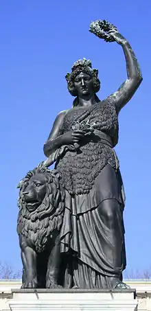 Farbige Untersicht auf eine weibliche Figur in griechischem Stil mit einem Löwen an der linken Seite. Die Figur drückt mit ihrer rechten Hand ein Schwert an ihre Brust und hält mit ihrer linken Hand einen Lorbeerkranz über ihr Haupt.
