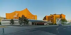 Frontale Farbfotografie von einem Gebäudekomplex im Sonnenuntergang. Das vordere flache Gebäude ist weiß mit einem breiten Eingang. Hinter dem Eingang sind zwei größere Gebäuden in Ockerfarbe mit wellenförmigen Dächern.