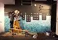 Hassani steht auf einer Bank und arbeitet an einem Graffiti das die ganze Wand einnimmt. Zu sehen ist eine Frau mit gelber Kleidung und hellem Kopftuch. Sie trägt Klaviertasten durch eine Stadtkulisse die Kopf steht.