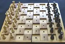 Ein aufgeklapptes Steckschachbrett von etwa Normgröße aus hellem und mittelbraunem, gemasertem Holz. Die schwarzen Felder sind gegenüber den weißen um etwa 5 mm erhöht. Die gedrechselten Holzfiguren sind für den Spielbeginn aufgebaut (links weiß, rechts schwarz), auf den Köpfen der schwarzen Figuren erkennt man dort eingelassene Schrauben.