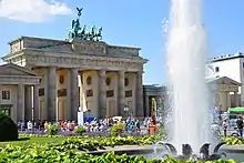 Blick auf Brandenburger Tor mit einer Grünfläche und Fontäne im Vordergrund. Das klassizistische Tor besteht aus sechs Säulen mit antiken Reliefszenen in Durchgängen, einem Giebelfeld mit antiker Szene und auf dem Dach die Quadriga in oxidiertem Grünspan.