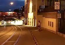 Eine lange Bahnunterführung in Augsburg mit leichtem Gefälle, glänzende Meterspur-Straßenbahnschienen mit etwa 1,50&nbsp;m Abstand vom Bordstein, Überholverbot (durchgezogene weiße Linie) für Autos in der beleuchteten Unterführung nahe dem Hauptbahnhof mit einem rot-weißen Zug oberhalb der leicht gebogenen Tunnelüberwölbung. Eine Fahrradfahrerin (vermutlich) auf dem etwa 2,50 Meter breiten Gehweg rechts, ein Bettler sitzt ein wenig weiter unten am Rand an der hohen Hauswand. Verkehrsschild Fussgänger mit Zusatzschild Fahrradfahren frei und Hinweistafel „Radfahren auf der Fahrbahn erlaubt“. Links auf der gegenüberliegenden Straßenseite trennt keine Wand mehr den Bürgersteig von der aufwärtsführenden Fahrbahn ab. Blaue Stunde am Abend mit Gebäude einer Brauerei rechts im Bild.