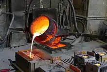 Aus einem rot glühenden Eimer, der an einer Stange befestigt und leicht nach vorne geneigt ist, fließt gelb glühende flüssige Bronze in ein Loch einer rechteckigen Form.