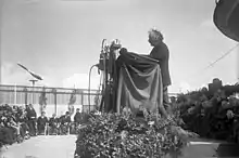 Schwarzweißfotografie von Einstein auf der Bühne in der Untersicht. Einstein steht im Seitenprofil zur Kamera und spricht von einem Rednerpult, das von Stoff umhüllt und mit vielen Mikrofonen ausgestattet ist. Das Publikum sitzt links und rechts sind Blumenarrangements.