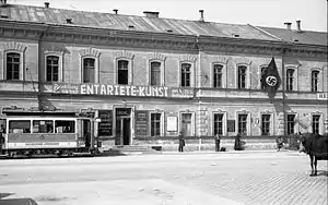 Frontale Schwarzweißfotografie von einer zweigeschossigen Häuserfassade mit dem Banner der Ausstellung „Entartete Kunst“.  Links fährt eine Straßenbahn und rechts stehen einige Menschen. Am Eingang befinden sich weitere Tafeln und eine Hakenkreuzflagge hängt am oberen, rechten Fenster.