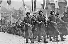 Frontale Schwarzweißfotografie von einer marschierenden Armee in langen Mänteln und Helmen. Ihr rechter Arm bewegt sich gleichzeitig nach vorne. Am oberen Bildrand hängen verschiedene Flaggen, darunter auch die Hakenkreuzflagge.