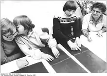 Das Schwarzweißfoto aus dem Jahre 1980 zeigt drei Jugendliche und eine erwachsene Frau an einem Tisch sitzend. Auf diesem liegen Bücher für Blinde, auf deren dunklen Seiten reliefartig, also tastbar, Tierdarstellungen (Löwe, Giraffe) eingeprägt sind. Links haben zwei Mädchen die Köpfe zusammengesteckt und tuscheln vergnügt miteinander. Die rechte der Beiden tastet dabei mit geschlossenen Augen mit den Fingerspitzen beider Hände über eine Buchseite, während sie der neben ihr Sitzenden ins Ohr flüstert. Diese, mit einer dicken Brille ausgestattet, hört belustigt zu. Der daneben sitzende, langhaarige Junge ist offenbar blind; er betastet konzentriert das vor ihm liegende Buch. Die Betreuerin schaut zu.