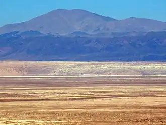 Die Cordillera Domeyko von den Hängen der Anden-Westkordillere aus gesehen. Auf 2.900 m ü. M. Höhe in 50 bis 70 km Entfernung in Richtung Westen betrachtet, erhebt sich hinter der Salar-de-Atacama-Senke (2.300 m ü. M.) und der Cordillera de la Sal der dunkle Steifen des El Bordo-Kliffs auf um die 3.000 m ü. M., vor dem höchsten Berg dem Cerro Quimal.