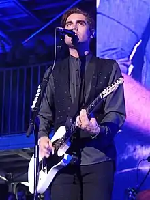Charlie Simpson singt auf einer Bühne in ein schwarzes Mikrofon hinein. Er trägt ein graues Hemd, eine schwarze Weste mit weißen Punkten und eine schwarze Hose. Er spielt auf einer schwarz-weißen Bassgitarre, die er um den Körper geschnallt trägt. Im Hintergrund sind eine Leinwand und mehrere Zuschauer auf dem Boden sowie auf einer Art Balkon stehend zu sehen.