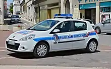 Foto eines Einsatzfahrzeugs der Police municipale Châteauneuf-sur-Charente, aufgenommen von schräg vorne, so dass Vorder- und Fahrerseite sichtbar sind. Der viertürige Pkw vom Typ Renault Clio III ist weiß lackiert und hat eine blaue Bauchbinde. Diese ist an der Fahrzeugseite im Bereich der Türen mit dem Schriftzug "Police municipale" in Versalien und blauen Lettern versehen. Auf der Fahrertür ist zudem das frankreichweit einheitliche Wappen der Police municipale angebracht, das dem Armabzeichen auf der Uniform entspricht. Auch auf der Fahrzeugvorderseite ist der Schriftzug "Police nationale" angebracht, in blau auf weißem Grund und eingefasst von einem roten Rechteck. Auf dem Dach des Autos ist ein Warnbalken mit Blaulicht installiert. Das Fahrzeug ist geparkt, offenbar auf einem Platz. Im Hintergrund befindet sich eine Straße mit Ladengeschäften.