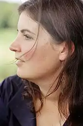 Auf dem Porträt blickt Christa Pfafferott nach links, sie ist im Profil zu sehen. Sie trägt eine dunkelblaue Bluse. Eine dunkelbraune Strähne ihres offenen, mittellangen Haares wird ihr vom Wind ins Gesicht geblasen.