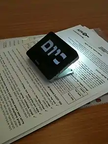Auf einem Tisch liegen einige Blätter mit hebräischem Text, darauf wurde das etwa zigarettenschachtelgroße Lesegerät platziert. Mit einer kräftigen Lichtquelle beleuchtet es einen kleinen Teil des Textes, auf seiner Oberseite erscheinen wenige hebräische Schriftzeichen in sehr starker Vergrößerung (mehrere Zentimeter hoch).