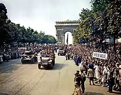 Frontale Farbfotografie in der Obersicht von einer breiten Allee mit Bäumen und einer riesigen Menschenmenge an beiden Seiten. Mehrere Panzer und Militärfahrzeuge mit offenem Verdeck fahren durch die Straße, die im Hintergrund einen Triumphbogen und eine Frankreichflagge hat. Rechts vorne stehen Krankenschwestern und Polizisten, die die Menge zurückhalten. Auf einem Schild steht „Vive de Gaulle“.