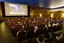 Ein voller Kinosaal in einem alten Programmkino. Viele Menschen sind von hinten auf ihren Plätzen zu sehen. Sie blicken nach vorn auf die Leinwand.