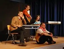 Die vier Mitglieder der Kabarettgruppe (drei Männer und eine Frau) bei einem Bühnenauftritt. Sie sitzen bzw. stehen um einen Tisch im Lehrerzimmer herum, den Bühnenhintergrund bildet ein dunkelblauer Vorhang. Einer singt zur Gitarre, ein zweiter hockt, bekleidet mit T-Shirt und Weste, an der Bühnenrampe und hat lässig ein rot-weiß gemustertes Palästinensertuch um den Hals geschlungen. Der dritte blickt – mit braun-kariertem Jackett und roter Kravatte deutlich erkennbar als eher konservativer Lehrer kostümiert – ratlos ins Publikum. Im Hintergrund steht ein Keyboard und wartet auf seinen Einsatz.