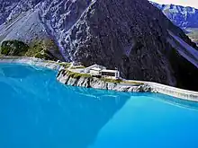 Blick aus Richtung einer Talsperre auf eine mehrgeschossige Berghütte, gebaut auf einem Felsvorsprung in Verlängerung der rechts daneben befindlichen Staumauer der Talsperre; im Hintergrund links ein Bergmassiv aus puren Fels; rechts der Talabgrund jenseits der Staumauer