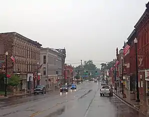 Eine breite, regennasse Straße mit hohen Geschäftsbauten auf beiden Seiten. Zwei sich annähernde Fahrzeuge haben die Scheinwerfer eingeschaltet. Im Bildhintergrund verengt sich die Straße an einer Brücke.