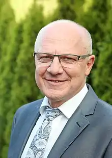 Portraitfoto des Gemeindeleiters Heinrich Klassen in einem grauen Anzug mit grau gemusterter Krawatte vor einer dunkelgrünen Hecke.
