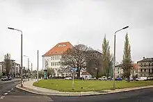 Foto eines vierstöckigen, breiten Gebäudes mit hohem Dach hinter einer Grünfläche. Auf der linken Seite des Gebäudes ist ein Fassadenbild zu sehen, das Rübezahl zeigt,